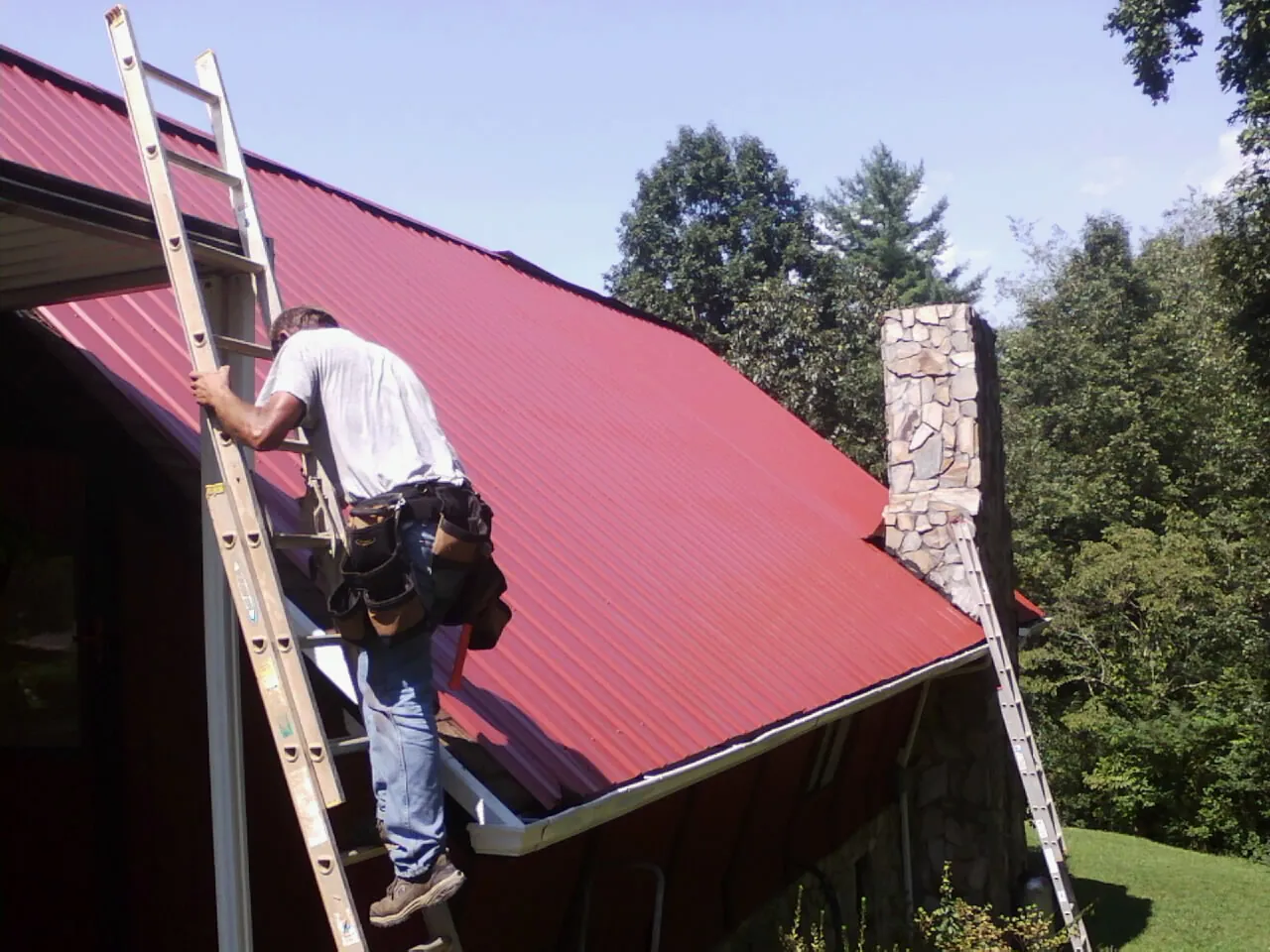 Roofer on a ladder beside a red metal roof and stone chimney — Soffit & Fascia Repair by Roofing Brothers in Prairie Du Sac
